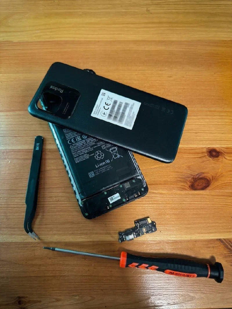 Móvil Redmi abierto con la batería a la vista, la tapa trasera encima, la placa de carga suelta debajo, unas pinzas curvas y un destornillador de precisión naranja sobre una mesa de madera.