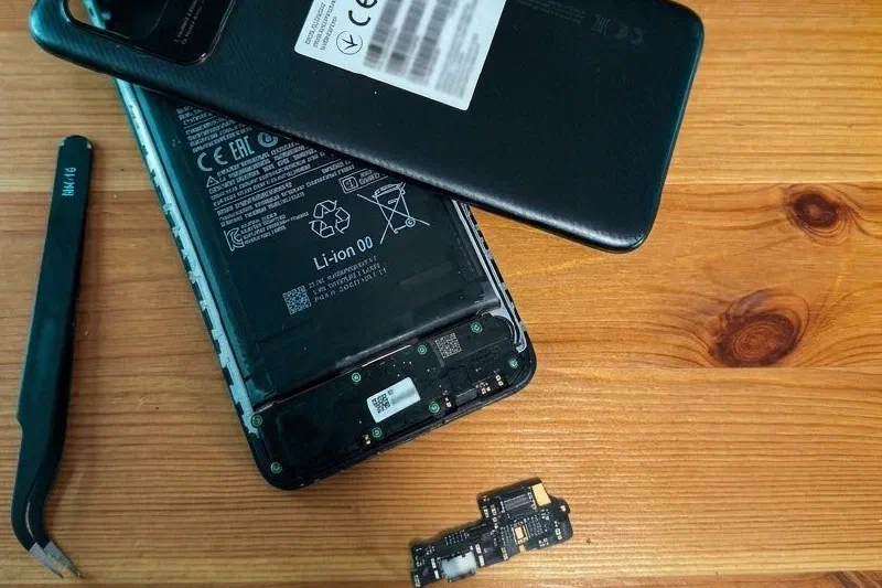 Móvil Redmi abierto sobre una mesa de madera, con la tapa trasera encima, la placa de carga suelta al lado, unas pinzas y un destornillador de precisión.