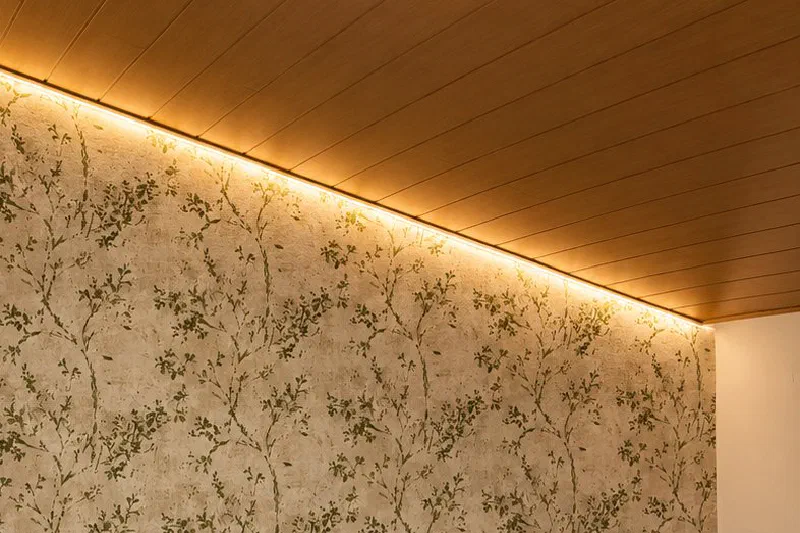 Pared empapelada con un estampado de ramas verdes sobre fondo beige, iluminada desde arriba por una tira LED cálida junto a un techo de madera.